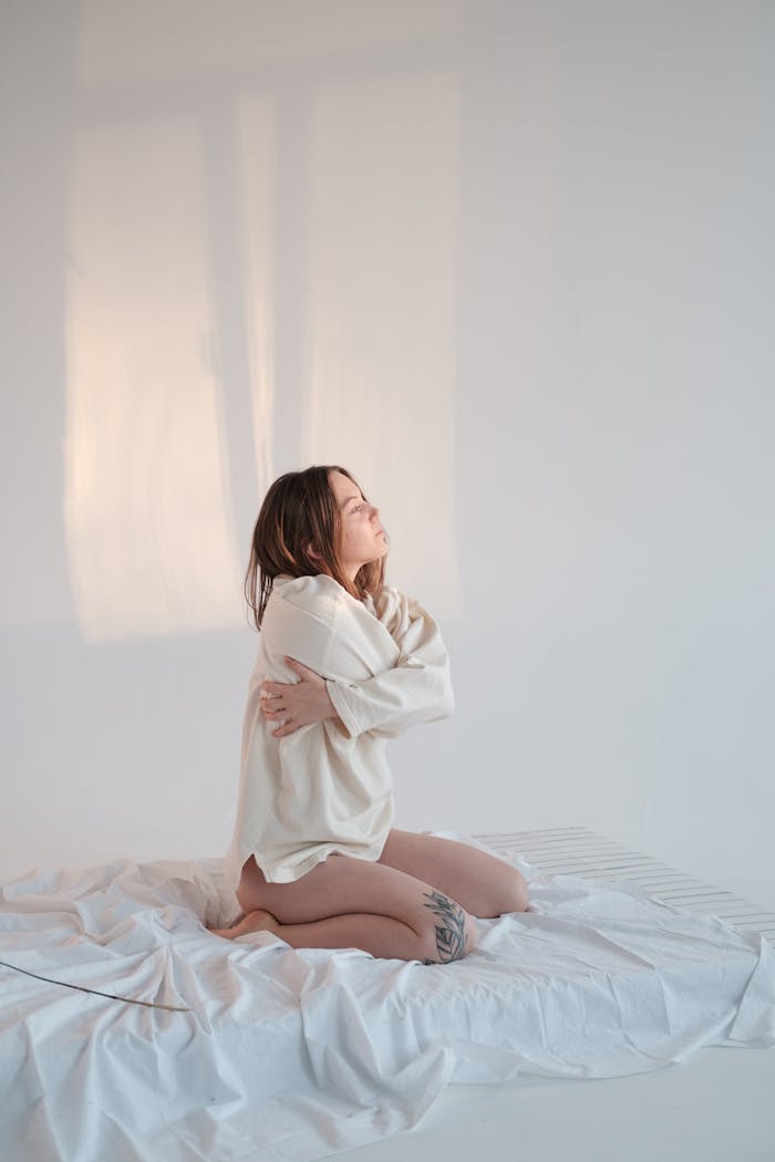 Woman kneeling on bed embraces herself, conveying solitude and introspection.